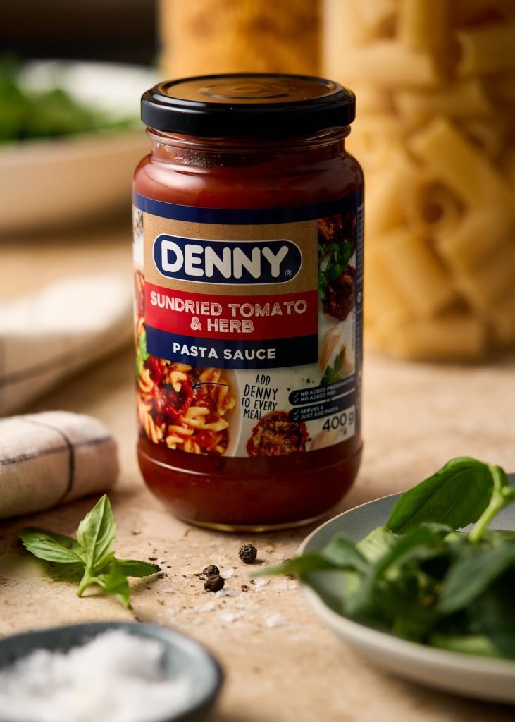 Hero shot of a jar of Denny Sundried Tomato and Herb pasta sauce on a warm stone surface with basil, peppercorns, flaky salt and dried pasta behind
