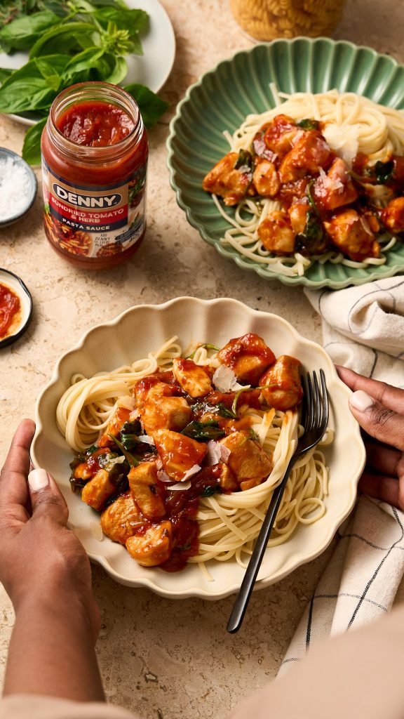 Hands placing a scalloped bowl of linguine with chicken in sundried tomato sauce on a warm stone surface, with a jar of Denny pasta sauce and fresh basil