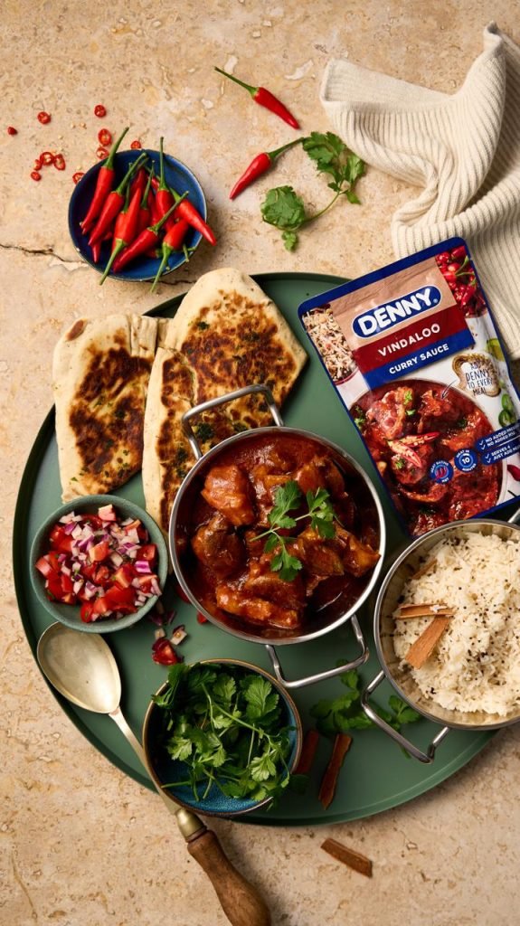 Overhead shot of a green tray set with vindaloo curry in a copper pot, naan, basmati rice, tomato salsa and coriander, with a Denny Vindaloo curry sauce sachet