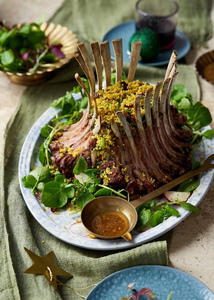 A crown roast of lamb filled with herbed couscous and dressed with watercress on a blue speckled platter, with a brass ladle of jus on olive green linen