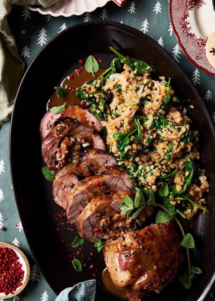 Overhead shot of sliced stuffed lamb loin with pink peppercorns, oregano and a lentil and spinach salad on a black oval platter over a Christmas tree print cloth