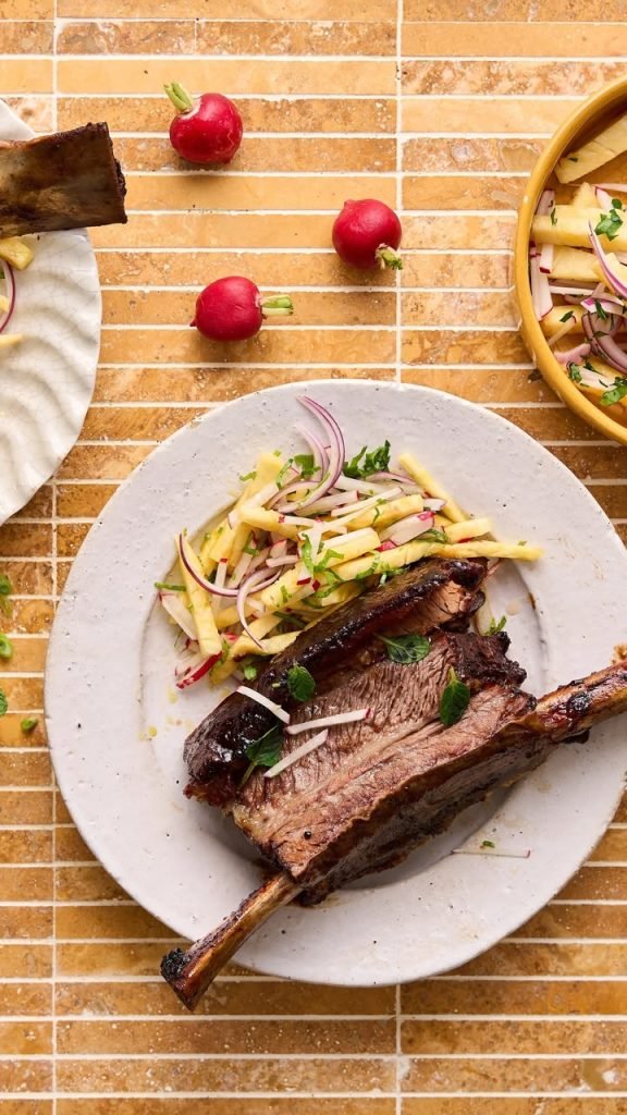 Overhead shot of sticky lamb ribs with apple and radish slaw and mint on a speckled ceramic plate over a honey-toned tiled surface