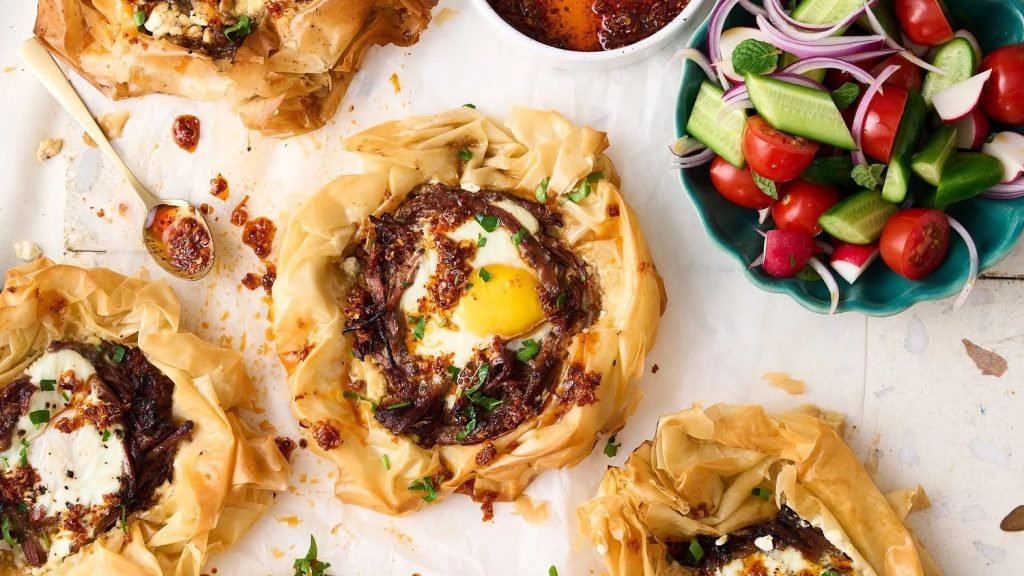 Overhead shot of crisp phyllo tarts filled with shredded beef, feta and baked egg, with chilli oil and a bowl of cucumber, tomato and red onion salad