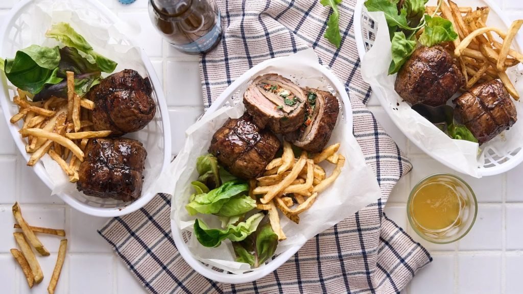 Overhead shot of three white baskets of stuffed rolled steak with skinny fries and butter lettuce on a navy checked cloth over white tiles