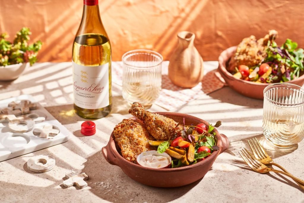 Crumbed chicken and salad in a terracotta dish with gold forks and ribbed glasses, beside a bottle of Leopard's Leap Unwooded Chardonnay against a warm plaster wall