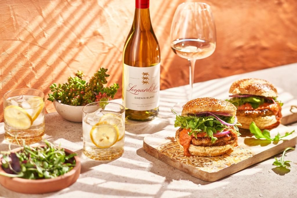 Two loaded burgers with rocket and red onion on a carved wooden board beside a bottle of Leopard's Leap Chenin Blanc, in dappled sunlight against a terracotta wall