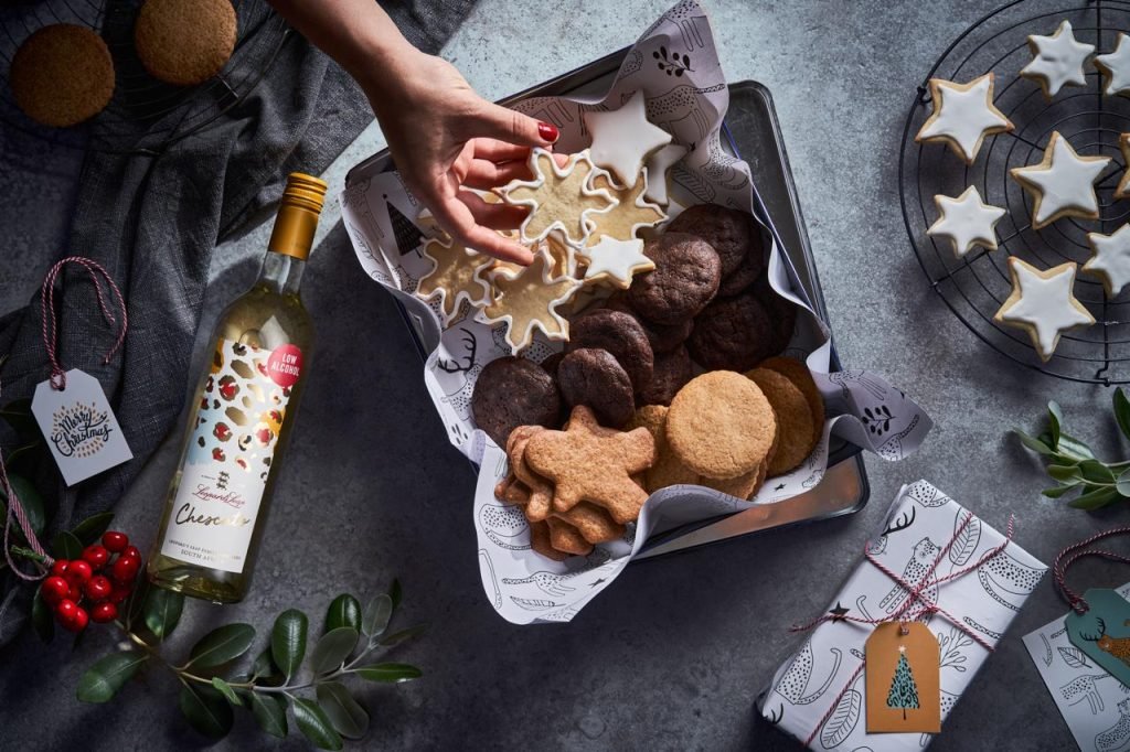 Overhead shot of a hand lifting an iced snowflake biscuit from a tin of assorted Christmas cookies, with a bottle of Leopard's Leap Chescato, gift tags and holly