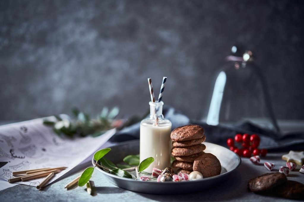 A stack of chocolate cookies and a bottle of milk with striped paper straws on a dark plate, styled with foliage and candy canes against a moody grey backdrop