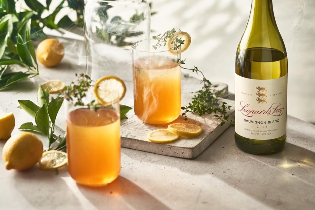 Two blush wine cocktails garnished with dehydrated lemon and thyme on a pale stone board, beside a bottle of Leopard's Leap Sauvignon Blanc in soft sunlight