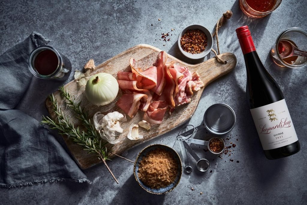 Overhead flatlay of streaky bacon, onion, garlic, rosemary and chilli flakes on a pale board against slate, with a bottle of Leopard's Leap Shiraz
