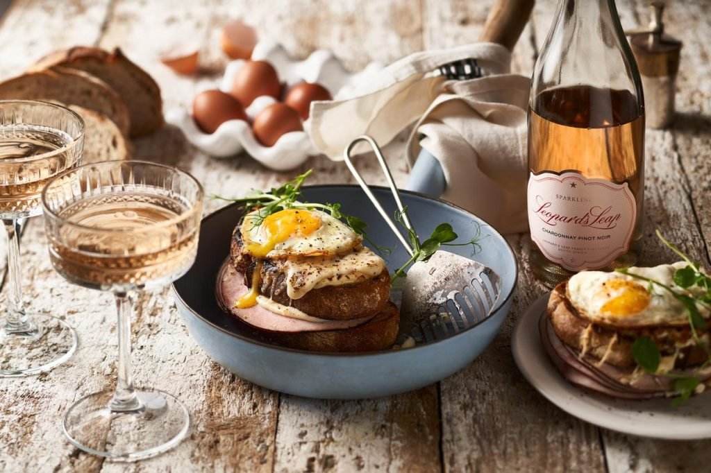 A croque madame with a runny fried egg in a blue bowl on weathered wood, with coupe glasses, brown eggs and a bottle of Leopard's Leap Sparkling Chardonnay Pinot Noir