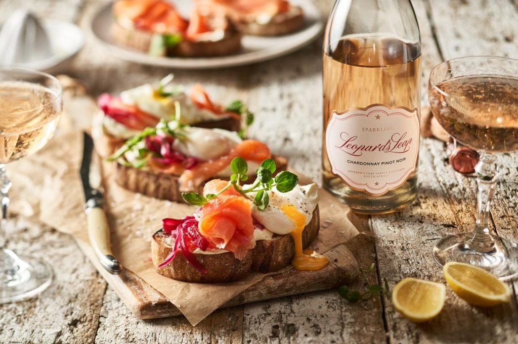 Sourdough toasts topped with smoked salmon, pickled beetroot and a runny fried egg on a rustic wooden table, with a bottle of Leopard's Leap Sparkling Rosé and coupe glasses