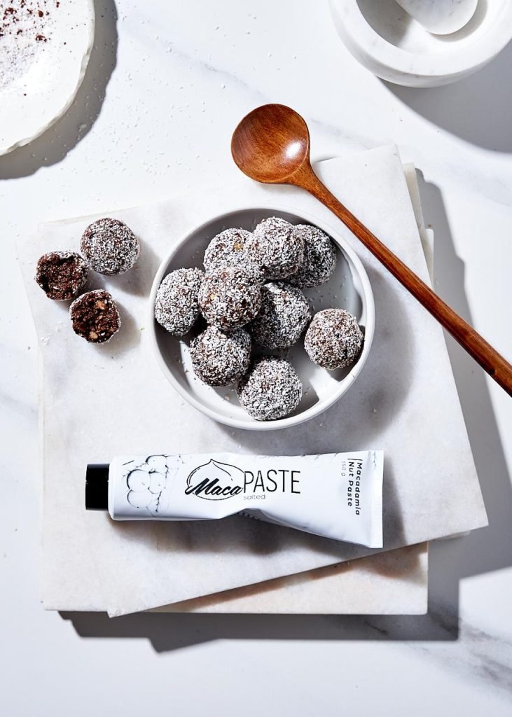 Overhead shot of cocoa and coconut bliss balls in a white dish on marble with a wooden spoon and a MacaPaste macadamia nut paste tube