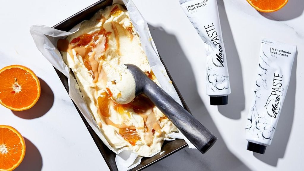 Overhead shot of a loaf tin of ice cream swirled with orange and macadamia caramel with a metal scoop, halved oranges and two MacaPaste tubes on white marble