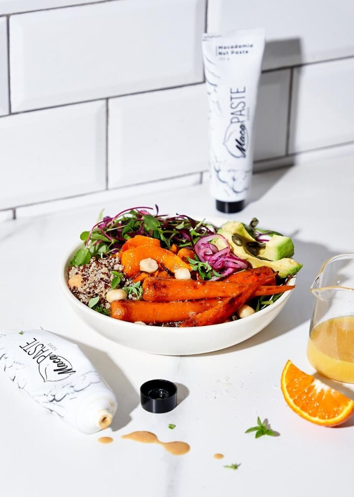 Roasted carrot, quinoa and avocado bowl against a white subway-tiled wall with a standing MacaPaste tube and a jug of orange macadamia dressing