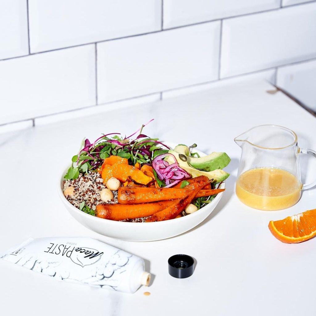 A white bowl of quinoa, roasted carrots, avocado, pickled onion and microgreens on a white counter, with a jug of orange dressing and a MacaPaste tube