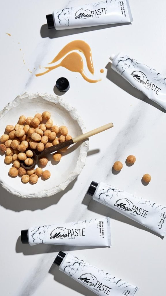 Overhead flatlay of five MacaPaste macadamia nut paste tubes on white marble with a swoosh of caramel-coloured paste and a stone bowl of roasted macadamia nuts