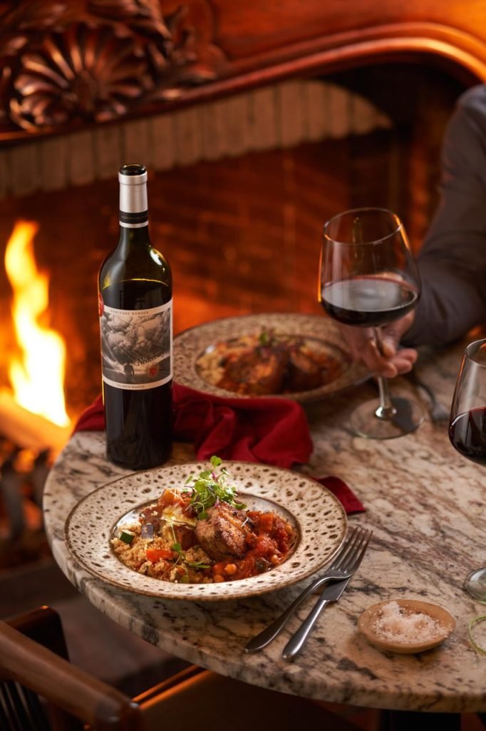 A plated lamb and couscous dish on a marble table beside a bottle of Nederburg Heritage Heroes red wine, with a hand raising a glass in front of an open fire