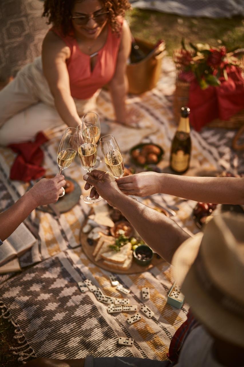 Overhead view of four hands clinking sparkling wine flutes over a woven picnic blanket set with a cheeseboard, dominoes and a flower-filled basket in golden light