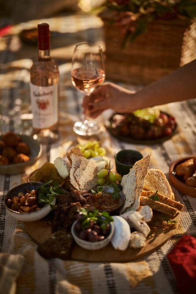 A generous picnic grazing board with cheese, crackers, olives, grapes and chutney, with a hand lifting a glass of rosé beside a bottle of Nederburg in golden light