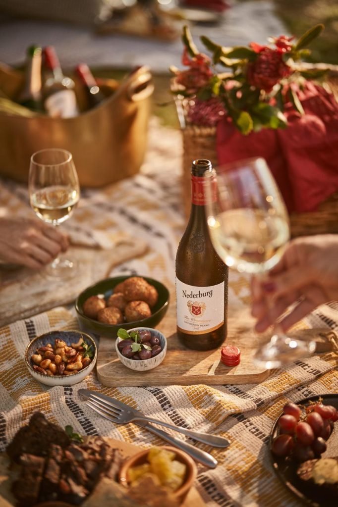 A chilled bottle of Nederburg Sauvignon Blanc on a wooden board with olives, spiced nuts and arancini on a woven picnic blanket in golden afternoon light