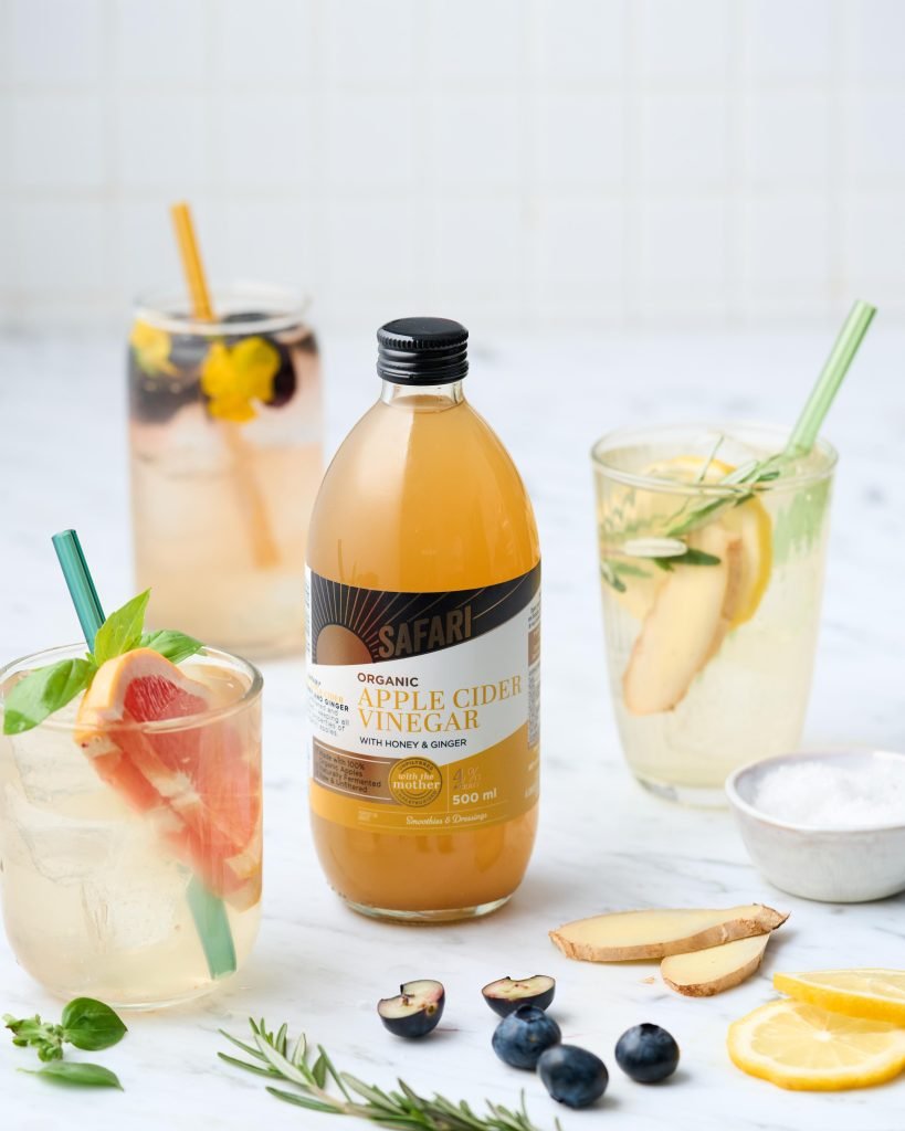 A bottle of Safari Organic Apple Cider Vinegar with honey and ginger, surrounded by three infused tonics with grapefruit, basil, ginger and lemon on white marble
