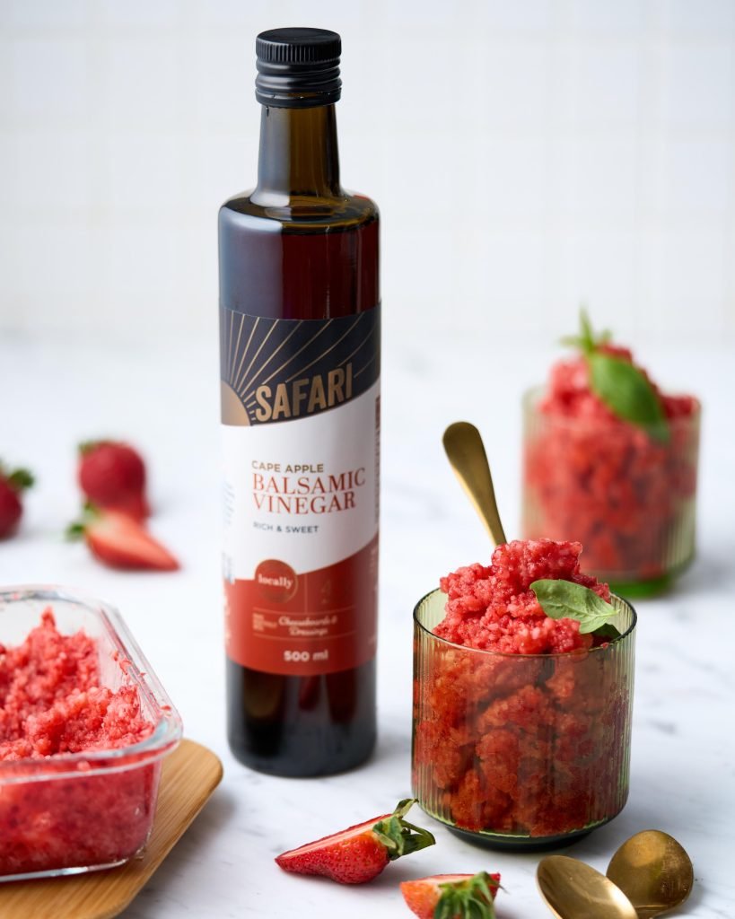 Ribbed glasses of bright red strawberry granita topped with basil and gold spoons, beside a bottle of Safari Cape Apple Balsamic Vinegar on white marble
