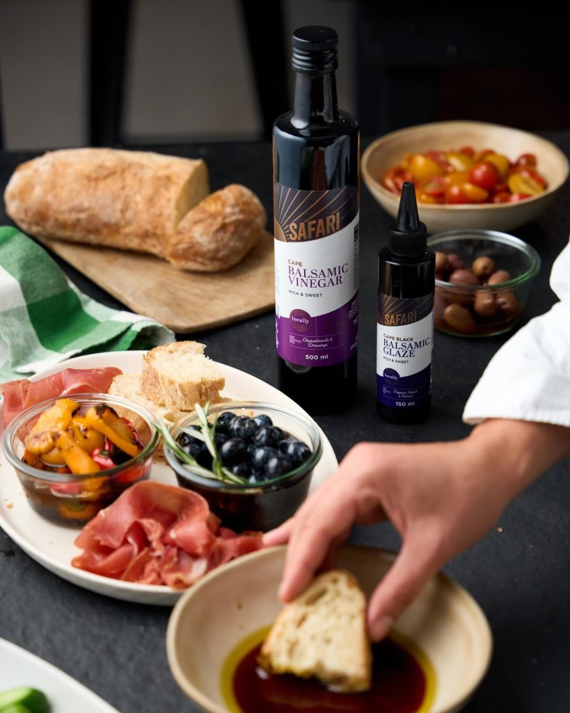 A hand dipping baguette into balsamic and olive oil beside a plate of prosciutto, marinated peppers and blueberries, with Safari Cape Balsamic Vinegar and Glaze bottles on a dark table