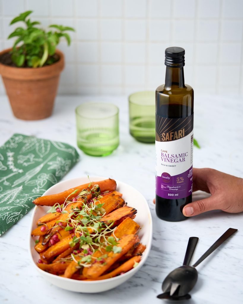 A white bowl of balsamic-roasted carrots topped with microgreens and pomegranate, with a hand placing a bottle of Safari Cape Balsamic Vinegar alongside