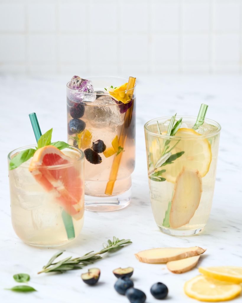 Three tall glasses of infused sparkling drinks with grapefruit, blueberries, rosemary, ginger and edible flowers on white marble against a tiled wall
