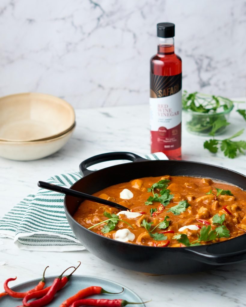 A black cast iron pan of butter chicken finished with yoghurt, coriander and red chilli on white marble, with a bottle of Safari Red Wine Vinegar behind