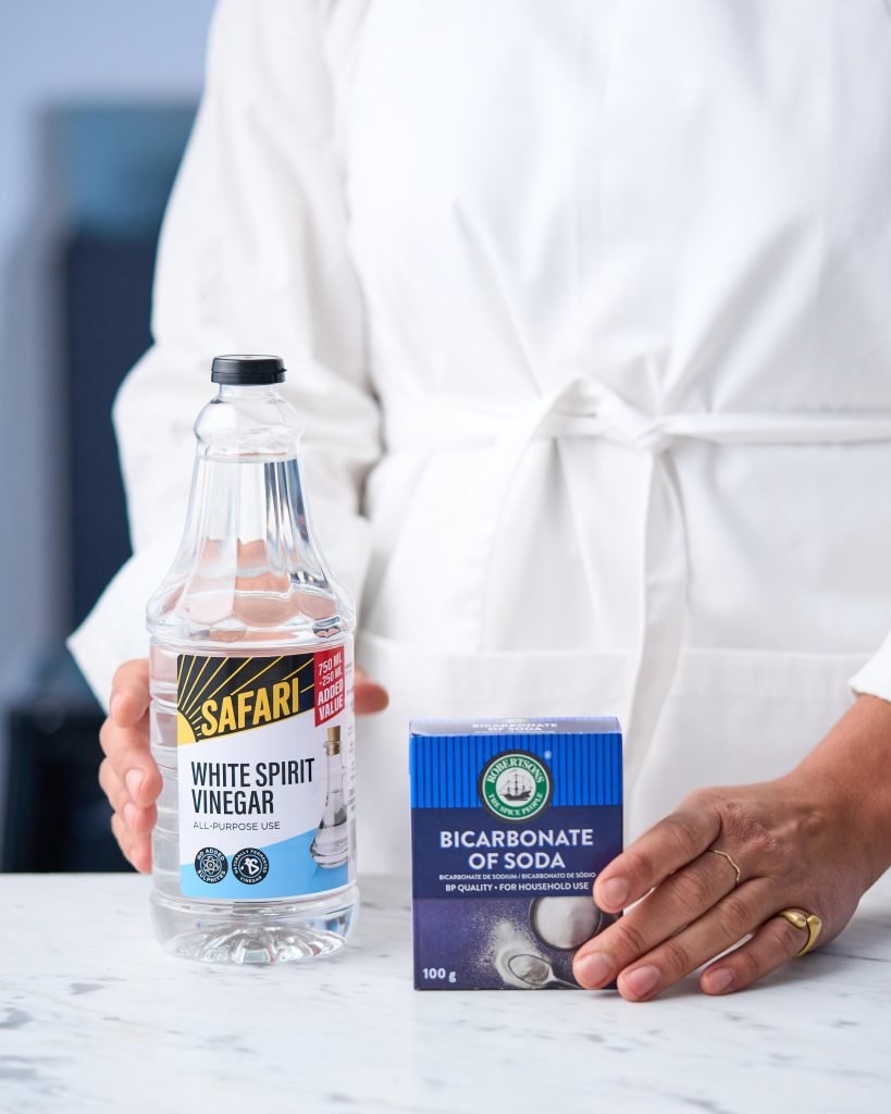 A person in a white apron holding a bottle of Safari White Spirit Vinegar and a box of bicarbonate of soda on a white marble counter
