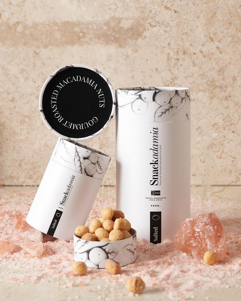 Two Snackadamia macadamia nut tubes with illustrated botanical packaging standing in crushed pink Himalayan salt against a travertine wall, with nuts in an open lid