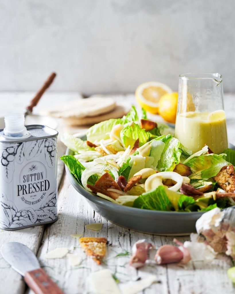 A grey bowl of caesar salad with fennel, shaved parmesan and croutons beside a jug of dressing and a Sutton Pressed macadamia oil tin on whitewashed wood