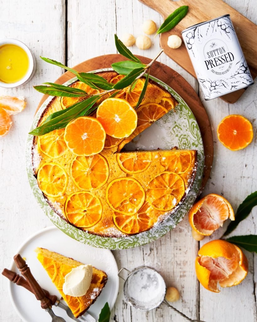 Overhead shot of a candied orange upside-down cake on a green patterned plate with fresh clementines, macadamia nuts and a Sutton Pressed oil tin on whitewashed wood