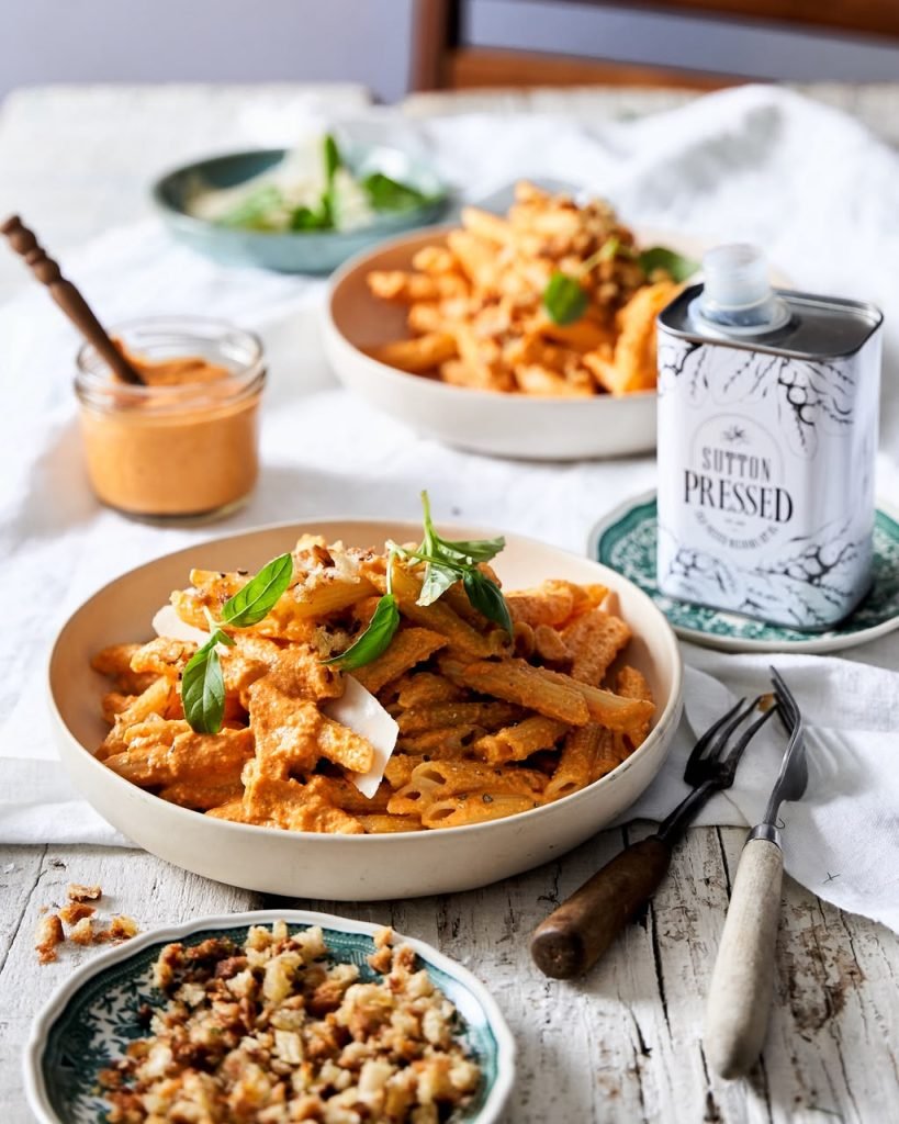 Two bowls of penne in romesco sauce topped with basil and shaved parmesan on a white cloth, with a jar of sauce and a Sutton Pressed macadamia oil tin