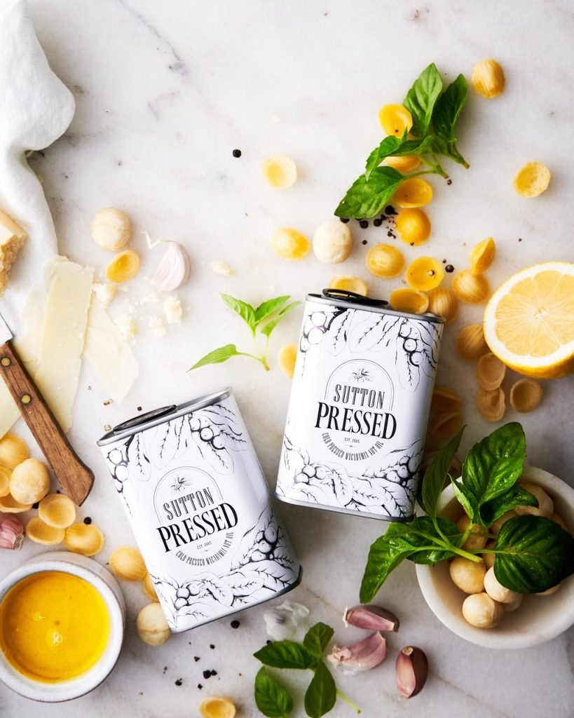 Overhead flatlay of two illustrated Sutton Pressed macadamia oil tins on marble with pasta shells, basil, garlic, lemon, parmesan and macadamia nuts